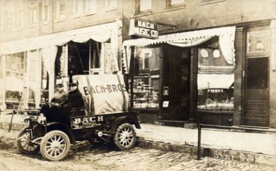 On an historic black and white but brownish photo you see a shop with a sign that tells the owner's name is Bach with a small miniature truck in front of the shop on the road. Two people are sitting.